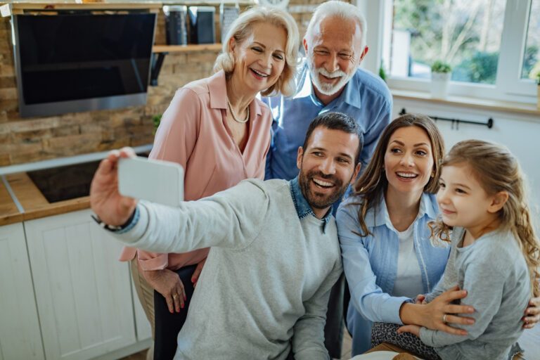 Família multigeracional sorridente, com avós, pais e uma criança, a tirar uma selfie em conjunto na cozinha de um apartamento.